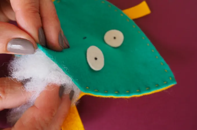 Hand adding white stuffing to felt soft toy with eyes on purple background.