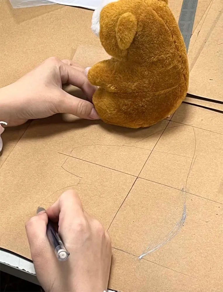 Hands sketching sewing pattern next to small brown plush toy on cardboard.