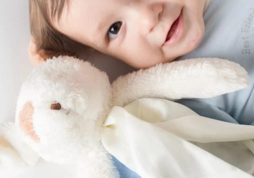 Baby Hugging Plush Bunny Smiling baby lying in bed cuddling a white plush bunny toy with a soft blanket